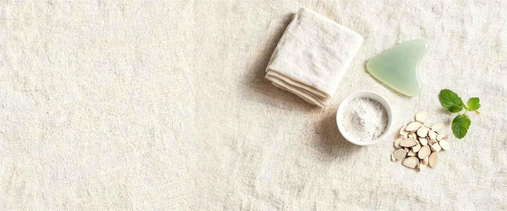 A jade gua sha stone, a bowl of pearl powder, dried bai zhi slices and a sprig of mint on cream linen.
